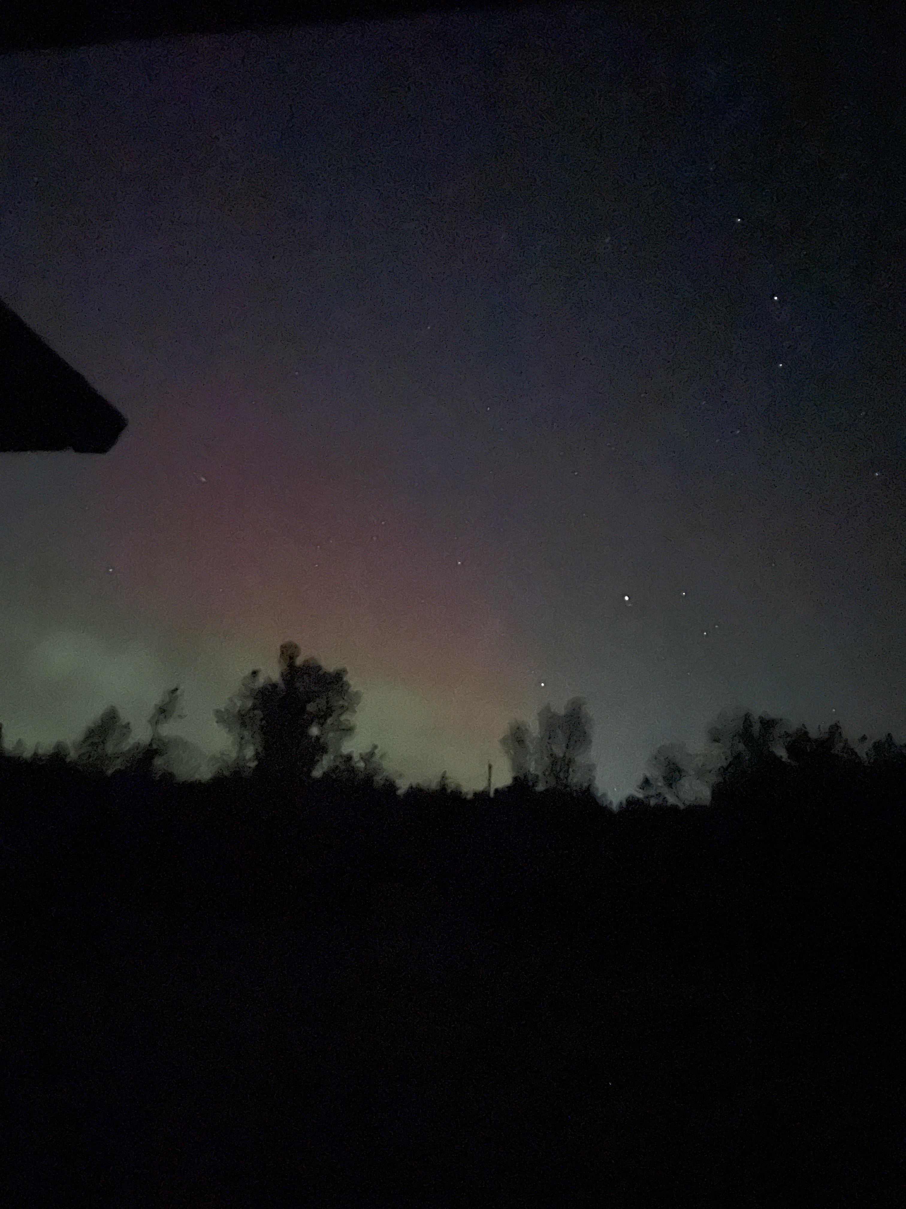 Night sky with stars and a rainbow-like phenomenon near the horizon, with tree silhouettes on the right.